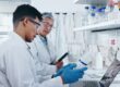 Two people wearing white lab coats sit at an open laptop. One is holding a bottle with a white label that they're both looking at.