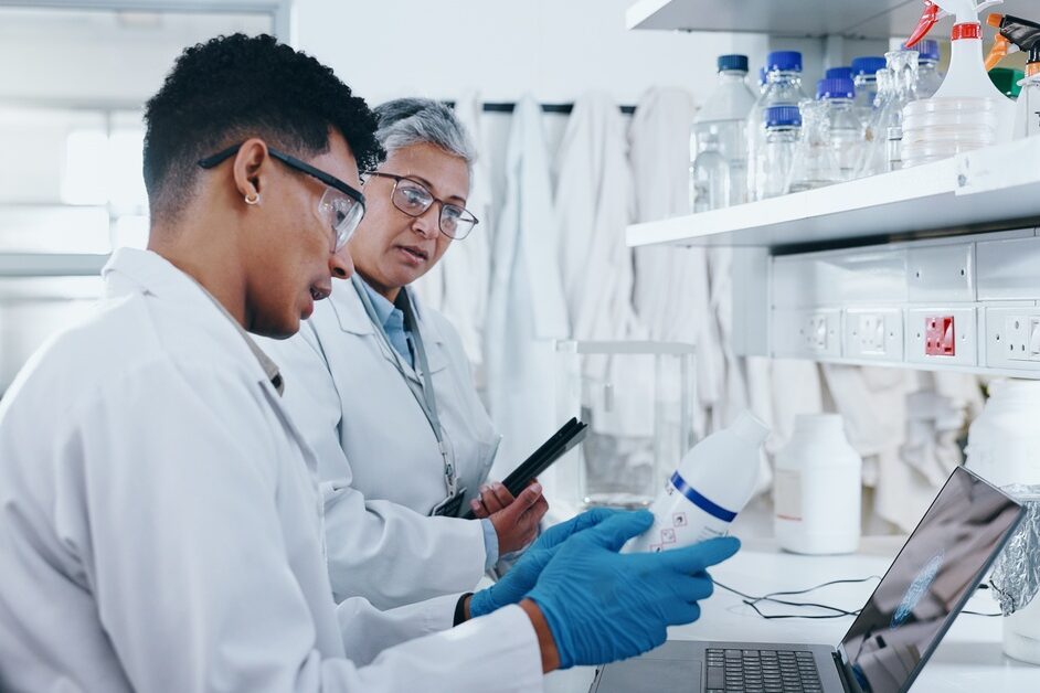 Two people wearing white lab coats sit at an open laptop. One is holding a bottle with a white label that they're both looking at.