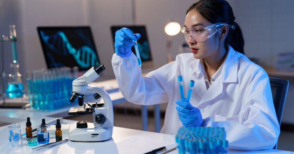 A woman wearing safety goggles and a white lab coat holds up a beaker with blue liquid in it. A microscope sits on a table.