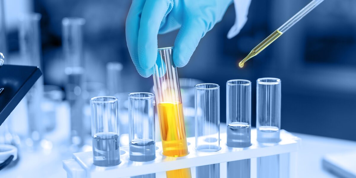 A person wearing blue gloves handles a beaker full of yellow liquid in a set of beakers. A pipette contains some of the liquid.