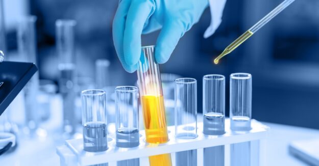 A person wearing blue gloves handles a beaker full of yellow liquid in a set of beakers. A pipette contains some of the liquid.