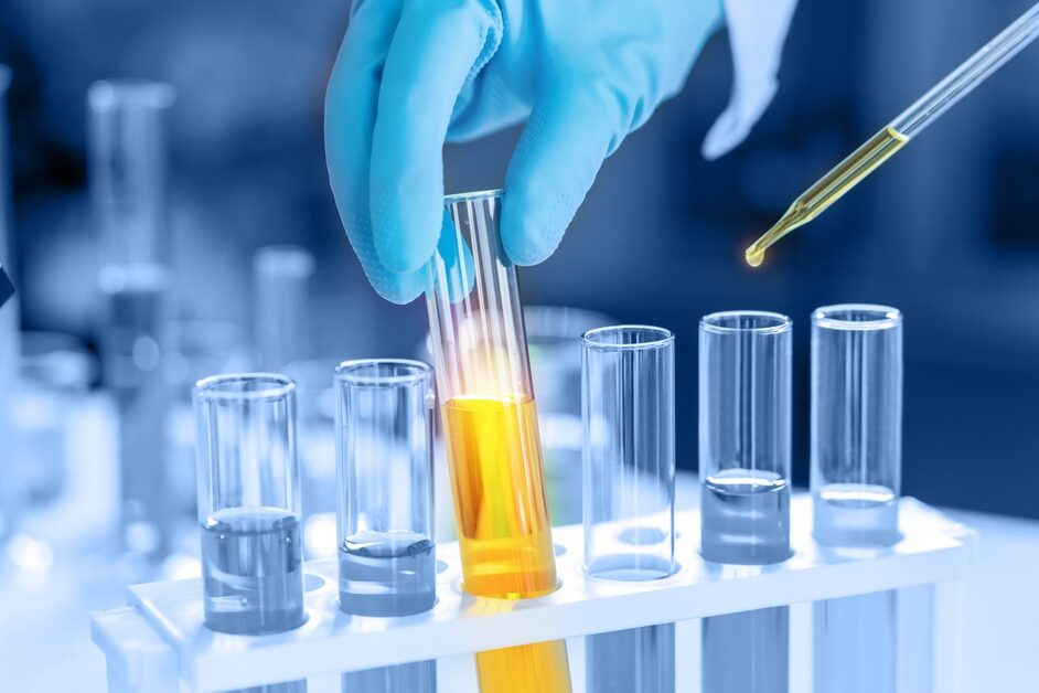 A person wearing blue gloves handles a beaker full of yellow liquid in a set of beakers. A pipette contains some of the liquid.