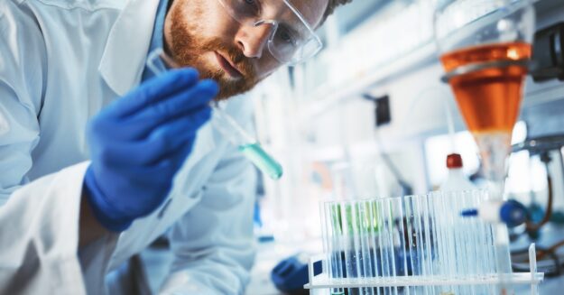 A man wearing safety goggles and a lab coat inspects a glass vial containing blue liquid in a laboratory.