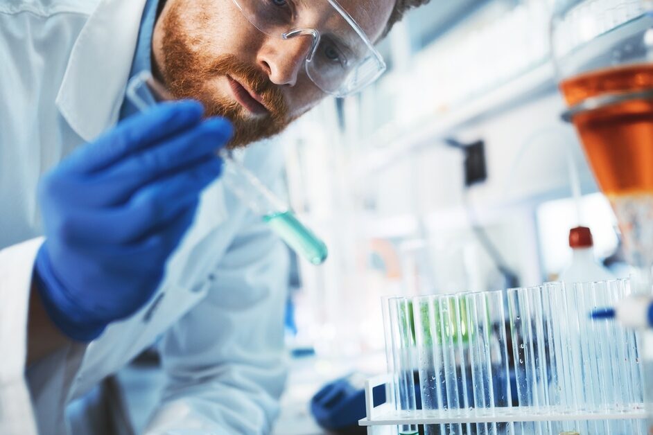 A man wearing safety goggles and a lab coat inspects a glass vial containing blue liquid in a laboratory.