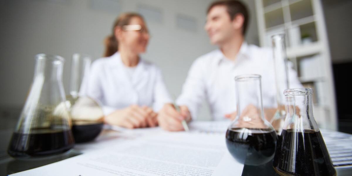 Two chemists review a contract at a lab table while glass flasks with dark liquid sit in the foreground.