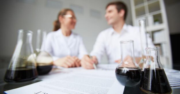 Two chemists review a contract at a lab table while glass flasks with dark liquid sit in the foreground.