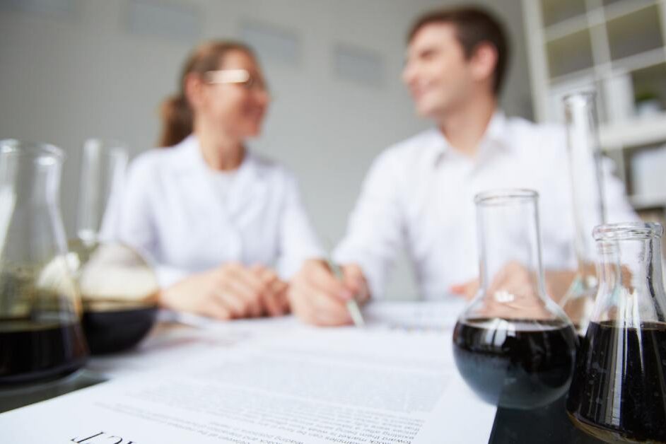 Two chemists review a contract at a lab table while glass flasks with dark liquid sit in the foreground.