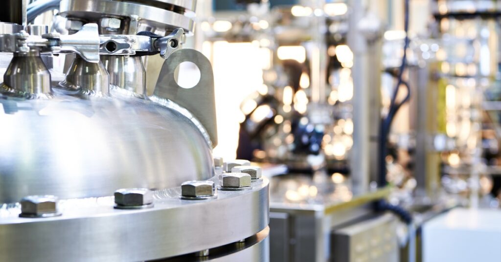  A stainless-steel chemical reactor with bolted fittings stands in an industrial processing facility with pipes in the background.