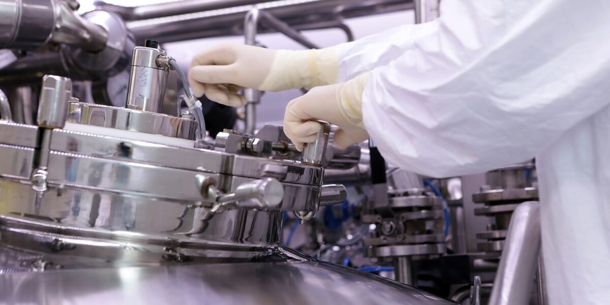 A technician in white protective clothing and gloves adjusts equipment on stainless-steel manufacturing machinery.