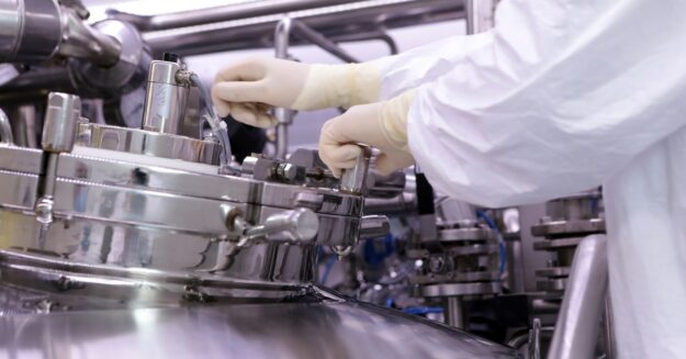 A technician in white protective clothing and gloves adjusts equipment on stainless-steel manufacturing machinery.