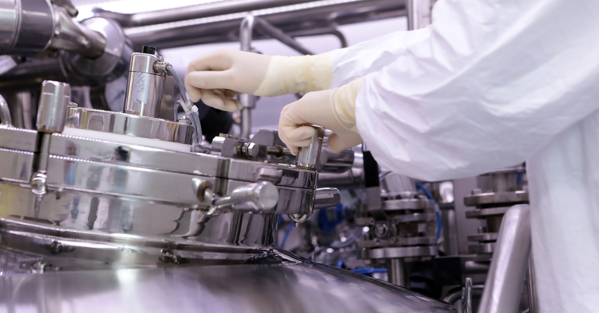 A technician in white protective clothing and gloves adjusts equipment on stainless-steel manufacturing machinery.