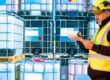 A man in a high-visibility vest is looking at a clipboard. He is also standing next to containers holding some liquid.