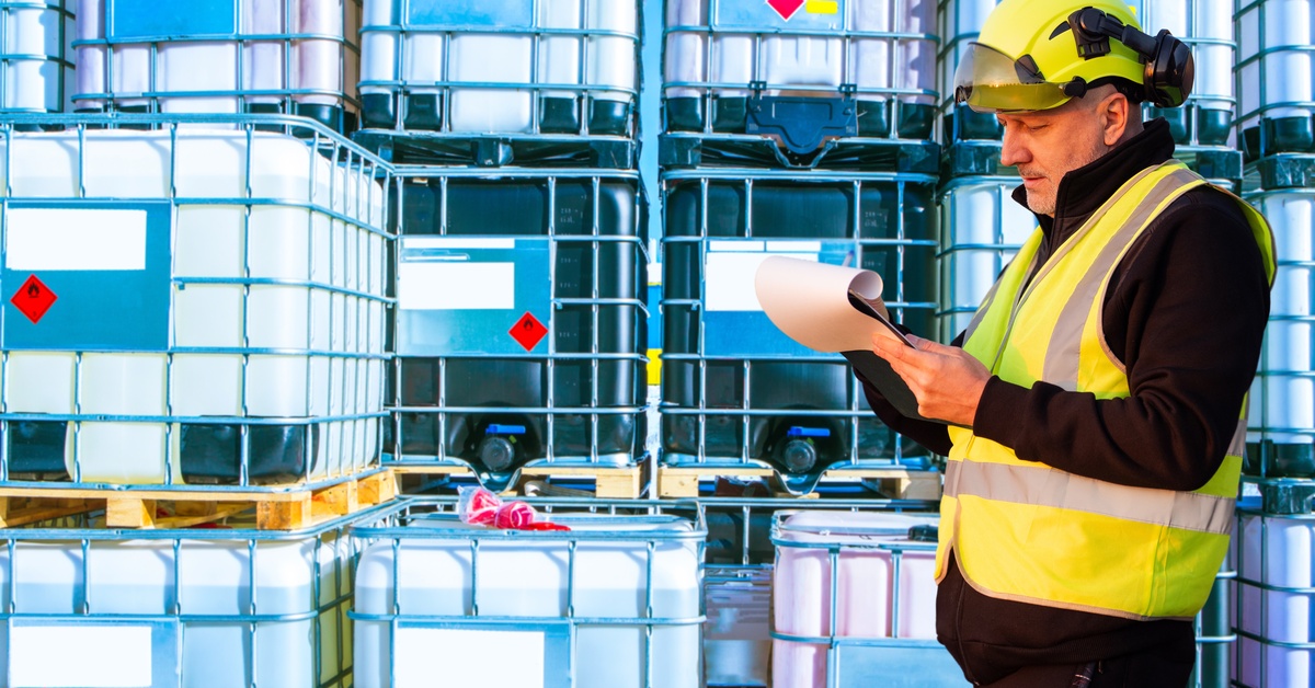 A man in a high-visibility vest is looking at a clipboard. He is also standing next to containers holding some liquid.