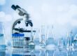 A microscope and laboratory glassware containing clear liquid sit on a reflective surface against a soft blue background.