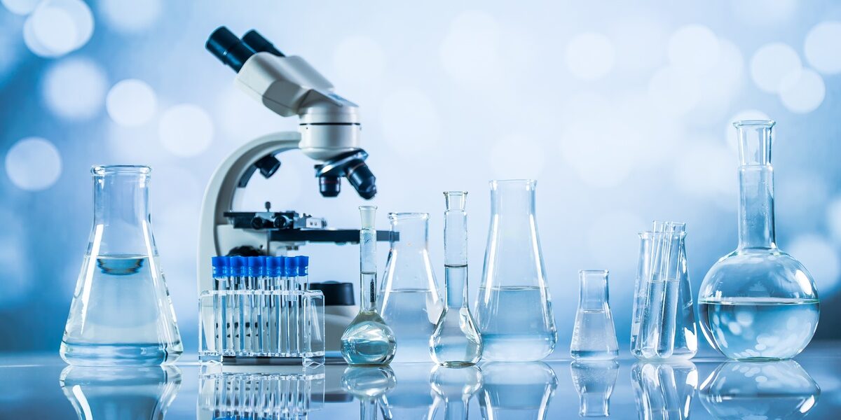 A microscope and laboratory glassware containing clear liquid sit on a reflective surface against a soft blue background.