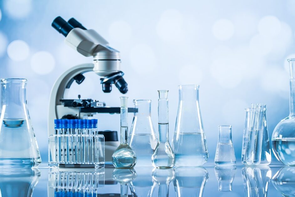 A microscope and laboratory glassware containing clear liquid sit on a reflective surface against a soft blue background.