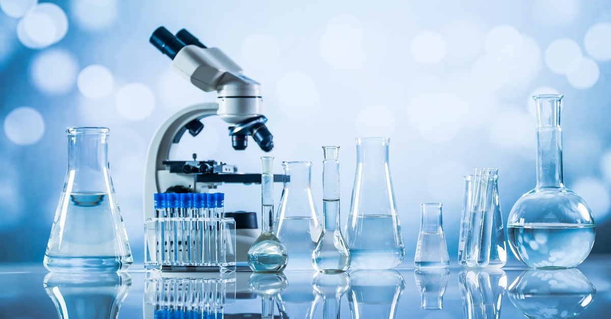 A microscope and laboratory glassware containing clear liquid sit on a reflective surface against a soft blue background.