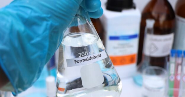 A person who is wearing a glove on their hand is holding a beaker filled with a clear liquid.