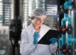 A woman in a white lab coat is holding a clipboard in one hand and a pencil in the other. There is machinery behind her.