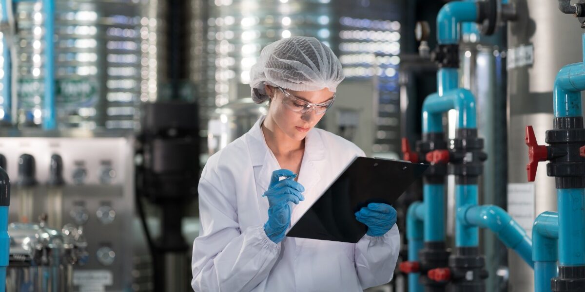 A woman in a white lab coat is holding a clipboard in one hand and a pencil in the other. There is machinery behind her.