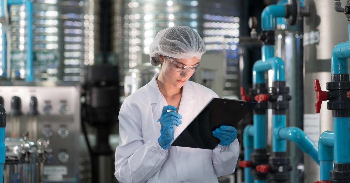 A woman in a white lab coat is holding a clipboard in one hand and a pencil in the other. There is machinery behind her.