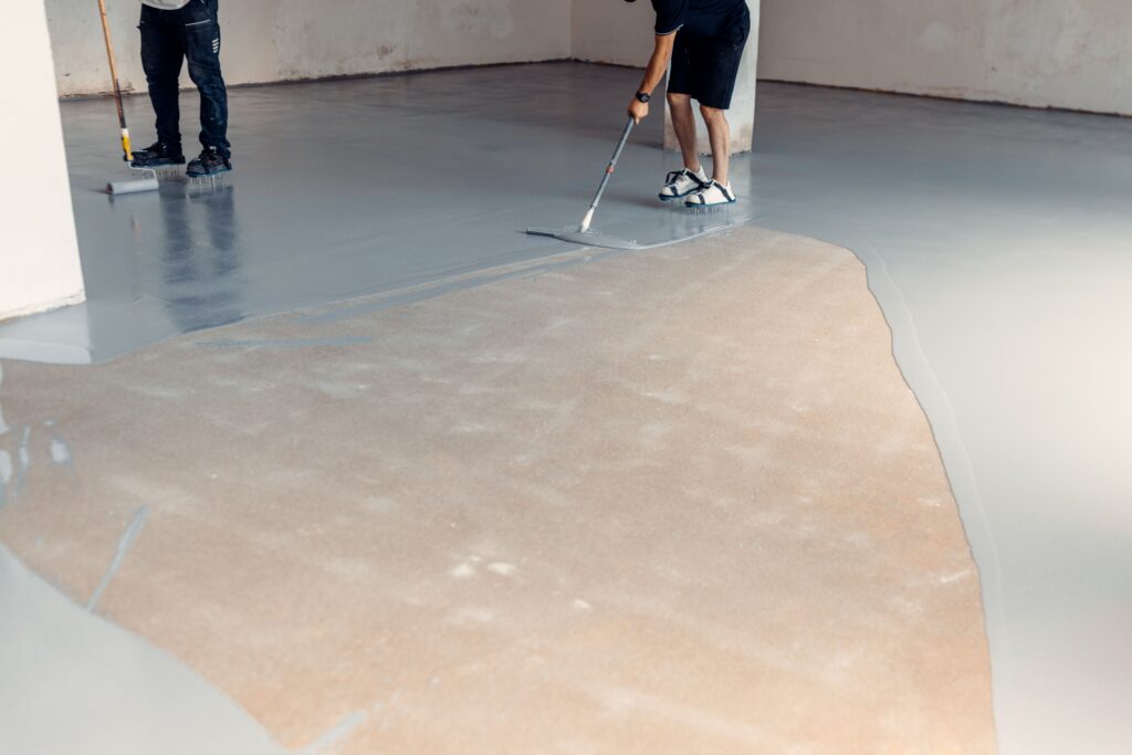 Two people are spreading an epoxy coating on a concrete floor. The epoxy substance is gray an glossy.
