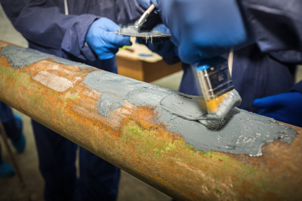 A person wearing blue gloves is covering a metal pipe in a substance. They are using a paint brush to apply the substance.