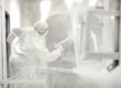 A person in a white jumpsuit is spraying down a metal piece that is suspended from the ceiling. There are other hanging pieces.