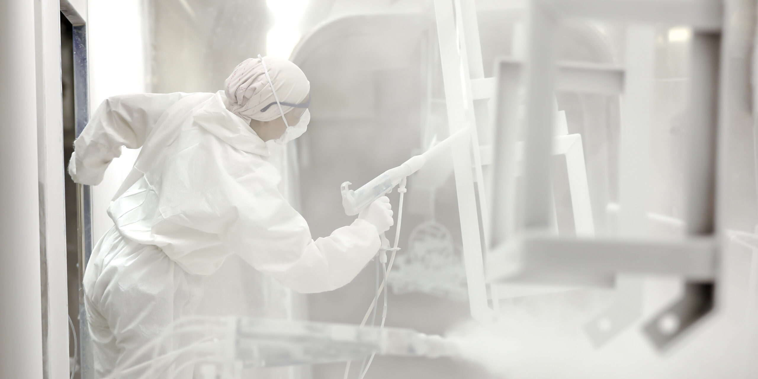 A person in a white jumpsuit is spraying down a metal piece that is suspended from the ceiling. There are other hanging pieces. A person in a white jumpsuit is spraying down a metal piece that is suspended from the ceiling. There are other hanging pieces.