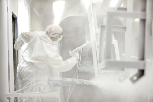 A person in a white jumpsuit is spraying down a metal piece that is suspended from the ceiling. There are other hanging pieces.