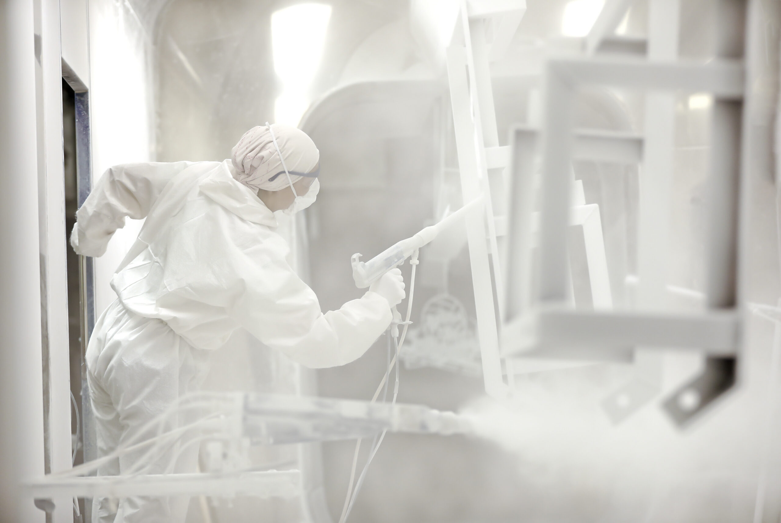 A person in a white jumpsuit is spraying down a metal piece that is suspended from the ceiling. There are other hanging pieces.