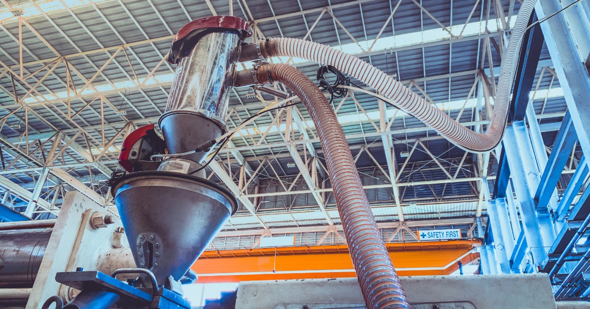 An industrial machine with a funnel has several tubes attaching to it. There is an orange walkway in the background.