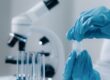 A person wearing blue gloves and a white protective shirt is handling a test tube with a liquid in it.