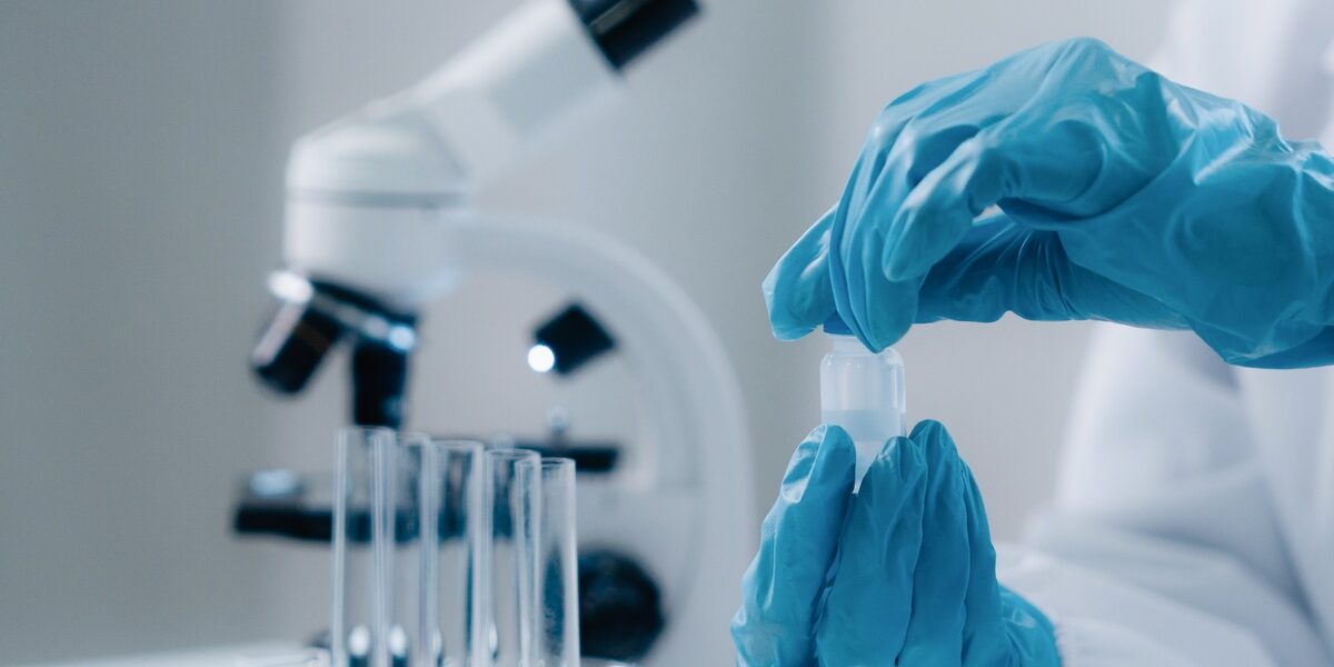 A person wearing blue gloves and a white protective shirt is handling a test tube with a liquid in it. A person wearing blue gloves and a white protective shirt is handling a test tube with a liquid in it.