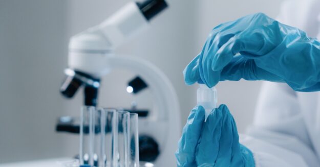 A person wearing blue gloves and a white protective shirt is handling a test tube with a liquid in it.