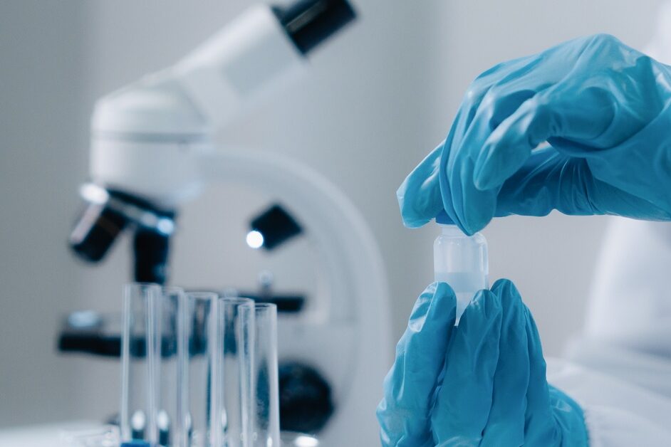 A person wearing blue gloves and a white protective shirt is handling a test tube with a liquid in it.