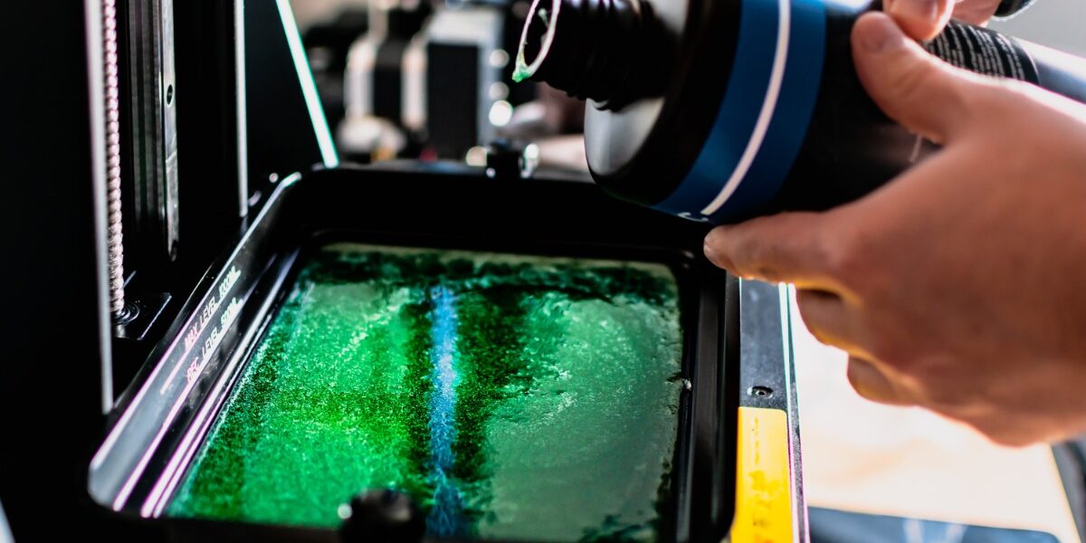 A person is holding a plastic container and pouring a solution into a larger container. There is a green liquid in the larger container.