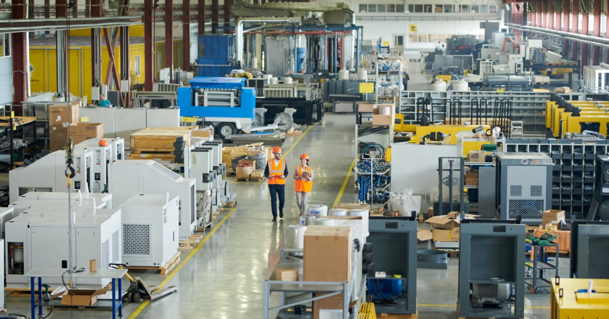 Two people are walking side by side while in an industrial looking warehouse. The people are wearing hi-vis gear.