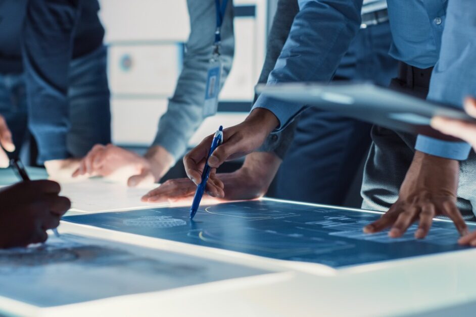 Several people are gathered around a table. There is a blue document on the table that everyone is looking at.