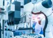 A robotic arm is reaching for a vial. The arm is in a sterile, industrial environment. There are red wires.