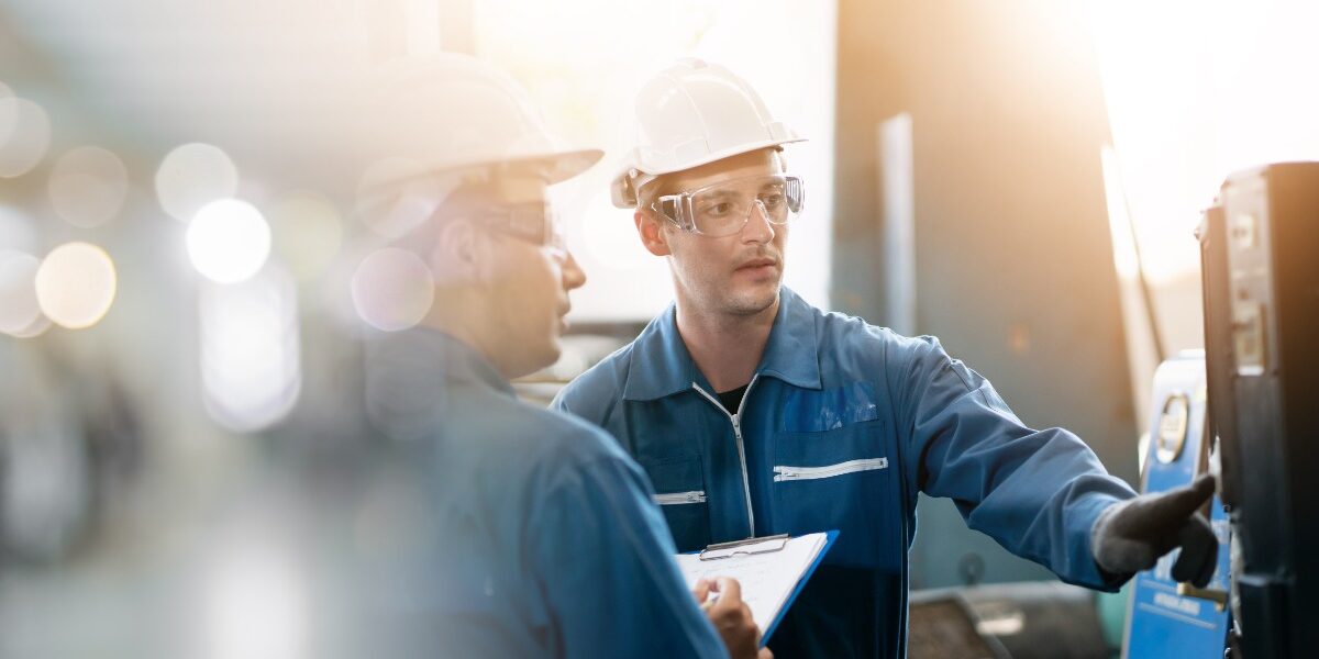 A man in a blue jumpsuit and a white hardhat is looking at a piece of machinery with another person next to him.
