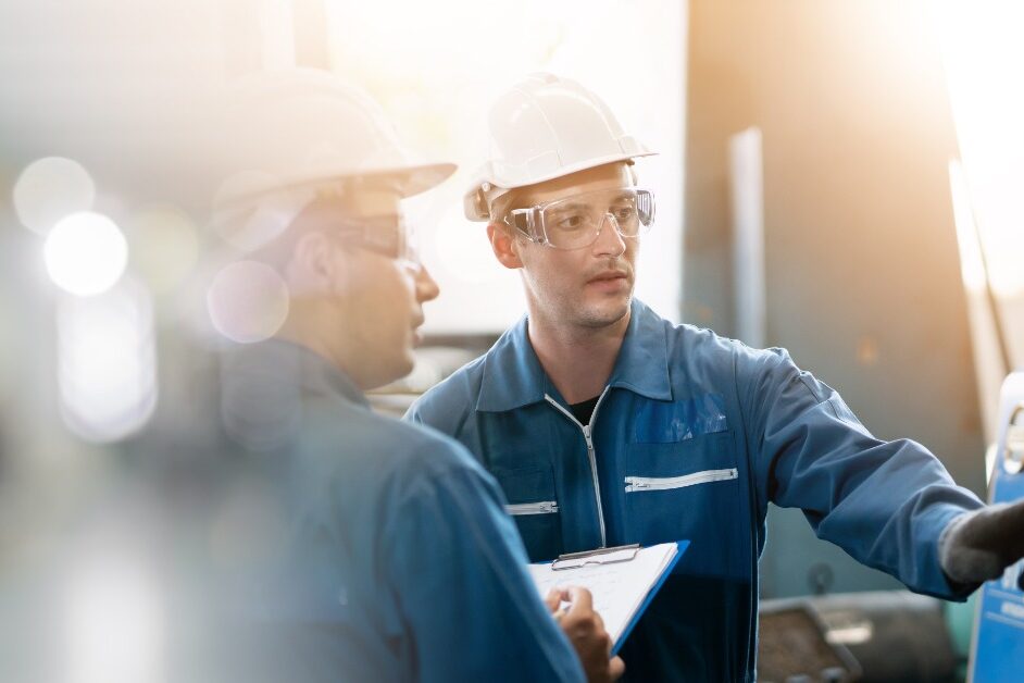 A man in a blue jumpsuit and a white hardhat is looking at a piece of machinery with another person next to him.