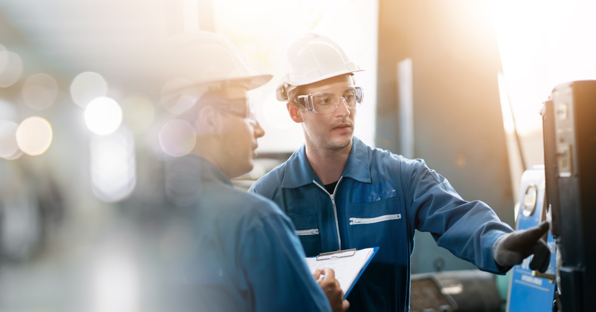 A man in a blue jumpsuit and a white hardhat is looking at a piece of machinery with another person next to him.