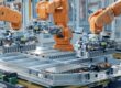There are several mechanical arms next to an assembly line. All the arms are painted orange.